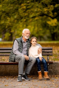Handsome grandfather spending time with his granddaughter on bench in park on autumn day