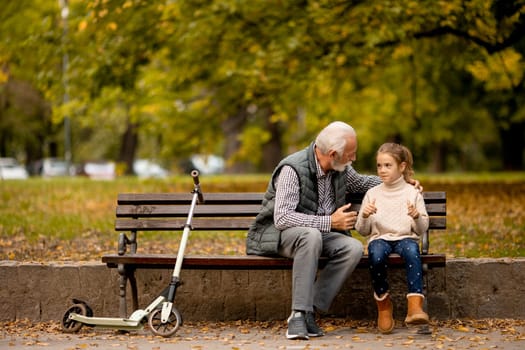 Handsome grandfather spending time with his granddaughter on bench in park on autumn day