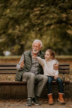 Handsome grandfather spending time with his granddaughter on bench in park on autumn day