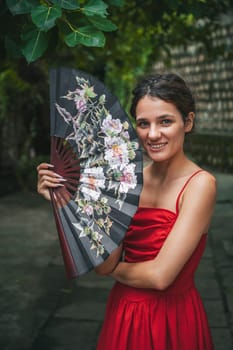 beautiful happy young woman in red dress with a fan in the garden. Tourism and travel in Asia