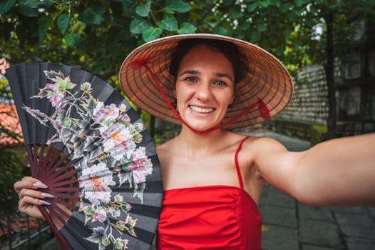beautiful young female tourist traveler with fan in a Vietnamese hat Non La takes a selfie in a garden. Tourism and travel in Vietnam