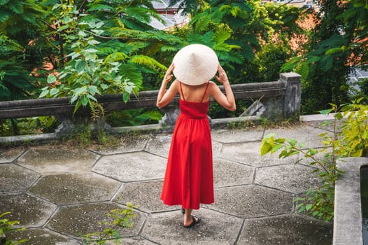 back of beautiful girl tourist traveler in a Vietnamese non la hat in red dress in a garden in Vietnam. Tourism and travel in Asia