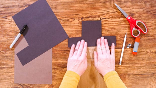 Step by step. Flat lay. Teacher guides online class through making a paper puppet from a brown bag, creatively using a wooden surface as a workspace.