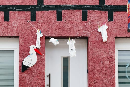 Beautiful view of baby clothes hanging on clothesline attached with clothespins and decorative stork figure in front of main door to house on spring day, side view medium shot. Concept street decor, national customs, travel time and tourism.