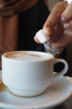A person carefully places a sugar cube into a steaming cup of coffee.