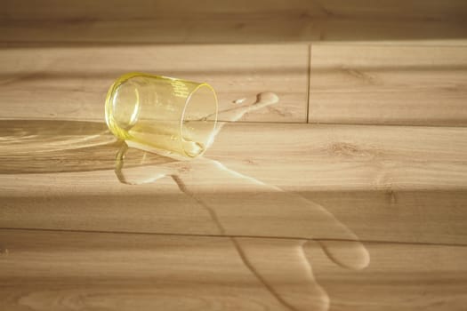 A yellow glass lies on a wooden floor with water spilling out, cast in sunlight.