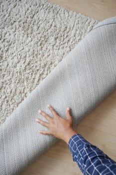 A hand is seen rolling up a gray rug on a wooden floor.