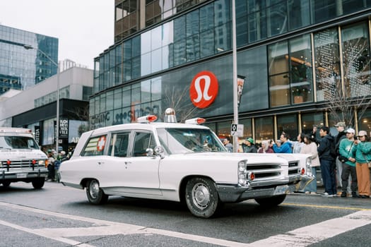 A vintage hearse is showcased on a parade route with a modern urban city backdrop