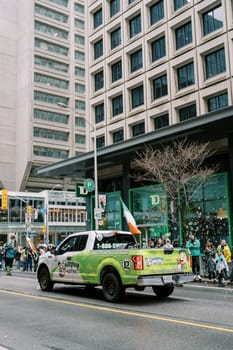 Colorful truck participating in a festive parade with flags and city backdrop.