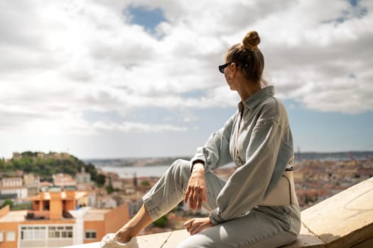 Young woman sitting on wall, enjoying the view of old town Lisbon. Tourist at Miradouro da Graca viewpoint, admiring scenic cityscape and architecture