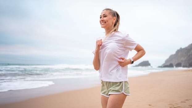 Woman jogging along sandy beach in morning. Athletic female in white top and green shorts running by ocean shore, enjoying nature and fitness outdoors