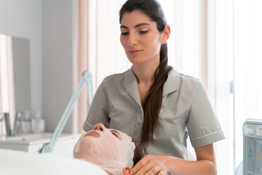 Professional masseuse fixing sheet mask on woman's face at cosmetology salon. Relaxed female customer is getting pampered in spa by beautician