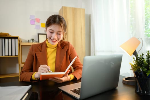 Smiling businesswoman writing in a notebook while working on her laptop at desk.