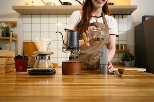Smiling woman in an apron pouring coffee beans into hand grinder, preparing fresh brew in a home kitchen.