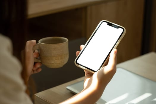 Young woman holding smartphone with blank screen and a ceramic coffee cup sitting in living room.