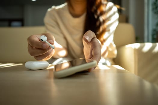 Young woman holding a wireless earbud and reaching for her smartphone.