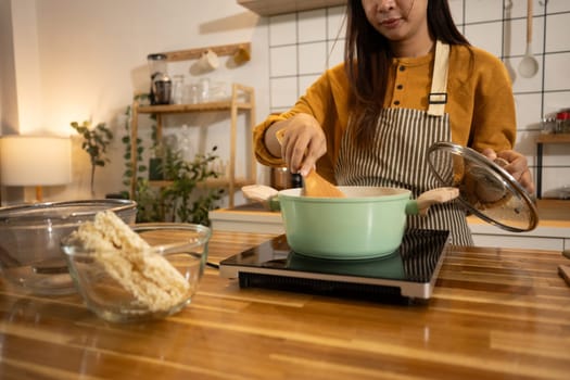 Young asian woman stirs pot on stove, preparing a homemade meal in a kitchen.