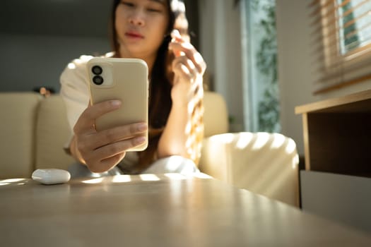 Young woman wearing a wireless earbud and using a smartphone in bright living room.