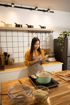 Smiling asian woman wearing an apron using smartphone while preparing meal in a kitchen.