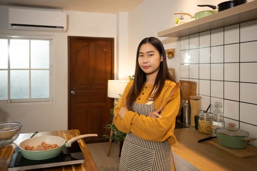 Proud young woman standing with arms crossed, holding a wooden spatula in a kitchen.