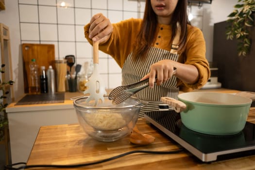 Woman wearing apron using kitchen utensils to transfer cooked noodles into a strainer.