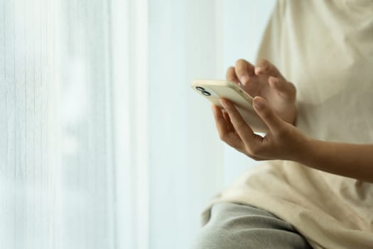 Young woman using a smartphone, sitting near a bright window with sheer curtains.