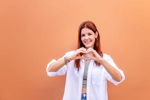 young woman standing in front of orange wall doing a heart shape with hands while looking at camera, she is wearing a white shirt, concept of love, copyspace for text