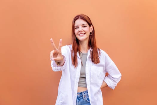 young woman standing in front of orange wall holding up two fingers in victory sign and smiling looking at camera, concept of peace, love and positivity, copyspace for text