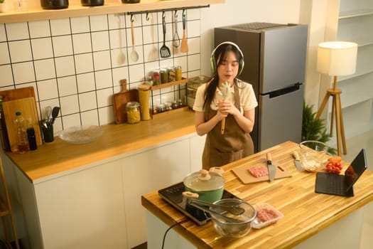 Happy young woman listening to music with headphones and enjoys cooking in a cozy kitchen.