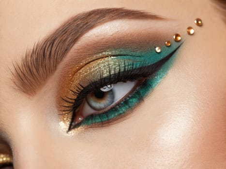 Close-up of a beautifully crafted eye makeup featuring shimmering gold and teal hues, enhanced with striking eyeliner. The intricate design is completed with decorative gems for added glamour.