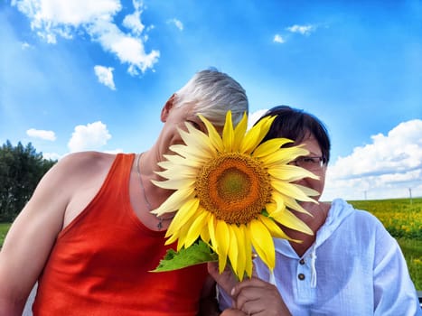 A middle-aged couple is having a joyful moment in the countryside, hiding their smiles behind a large sunflower while enjoying a sunny day during their travels.