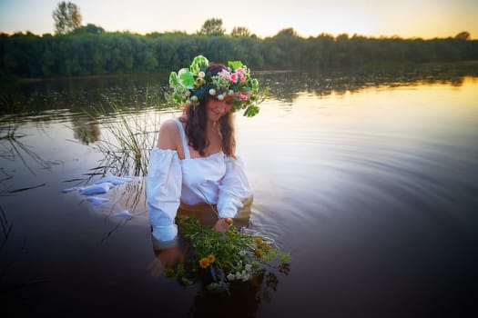 Slavic plump chubby girl in long white dress on the feast of Ivan Kupala with flowers and water in a river or lake on summer evening