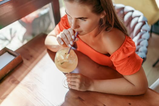 woman in a cafe seated, holding beverage. Dressed in an orange dress with her hair down. Bright interior, large windows allow natural light. Beverage consumption for refreshment