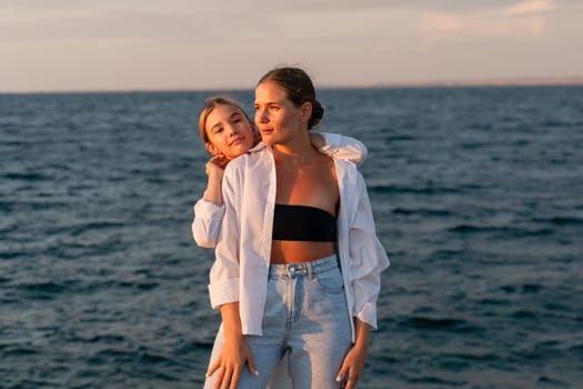 Two women, one in a white shirt and jeans, the other in a black bikini top, stand close together, looking towards the sea at sunset.