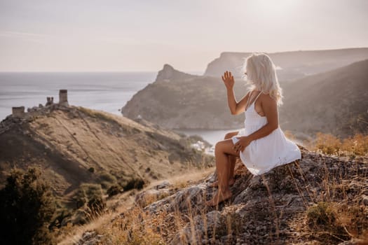 A woman is sitting on a hillside overlooking the ocean. She is wearing a white dress and has blonde hair. The scene is serene and peaceful, with the ocean in the background