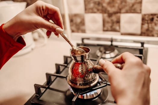 Close-up of hands stirring coffee in a cezve on a gas stove. The metallic cezve sits on a burner, its contents gently mixed.