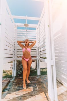 Woman in pink bikini rinsing off in an outdoor beach shower. White wooden structure, bright sunlight.