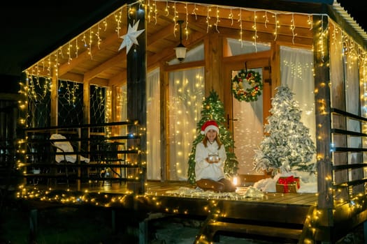 A decorated wooden cabin with a Christmas tree and string lights. A person wearing a Santa hat is seated on a bench, with a gift next to the tree.