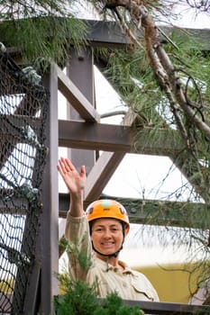 A woman wearing a helmet and khaki shirt smiles and waves from a wooden platform in a treetop obstacle course.