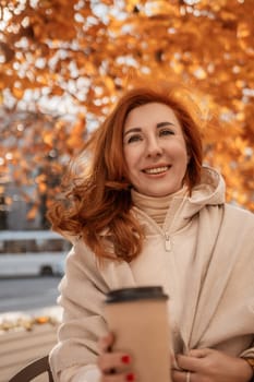 Smiling redhead woman, wrapped in a beige shawl, holds a coffee cup outdoors under a vibrant autumn tree. Golden leaves frame her.