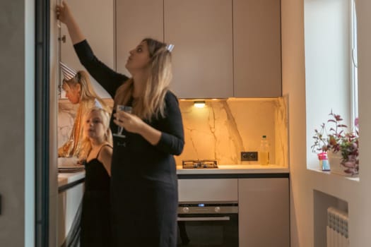 Woman in black dress reaches into kitchen cabinet, another woman holds wine glasses nearby. Modern, minimalist kitchen setting.