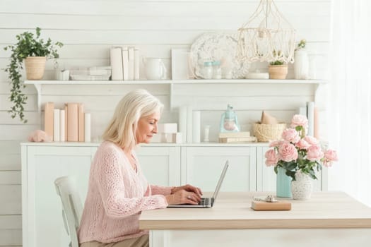 An elderly woman sits at a stylish wooden desk, focused on her laptop in a well-decorated home office. Soft pastel colors and a vase of roses enhance the serene atmosphere.