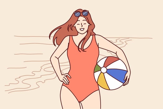 Woman in swimsuit with ball for beach volleyball stands on seashore enjoying summer vacation in tropical resort. Girl tourist smiles posing on beach near ocean and invites you to go on joint tour