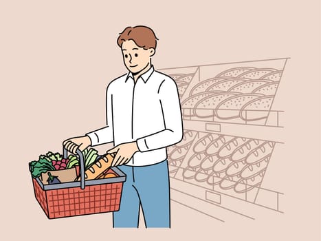 Man customer at grocery supermarket takes bread from shelf and puts it in basket of groceries. Guy visits supermarket or bakery buying fresh baguette and organic vegetables for preparing lunch.