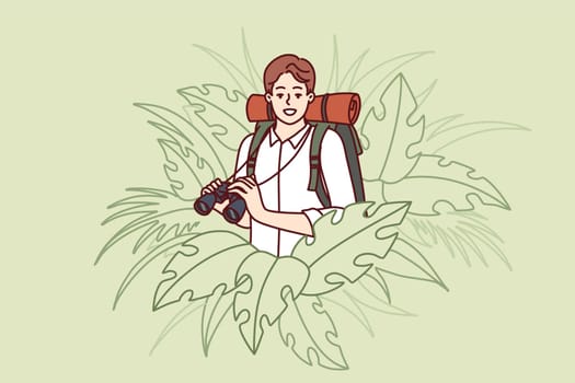 Man traveler stands in thickets of jungle and holds binoculars exploring area in search of further path. Guy traveler with backpack behind back loves adventure and is engaged in hiking
