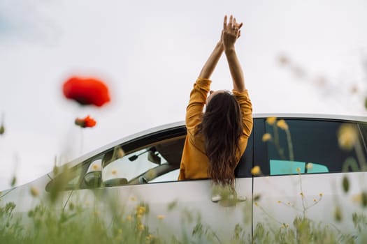 A woman is sitting in a car with her hands on her head, looking out the window. The car is parked in a field of flowers, with a red flower in the foreground. Concept of relaxation and freedom