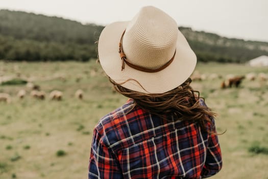 A woman wearing a straw hat and a red and blue plaid shirt stands in a field with a herd of sheep