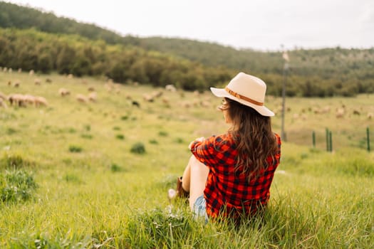 A woman wearing a straw hat and a red plaid shirt sits in a field of grass. The scene is peaceful and serene, with the woman looking out over the grassy field