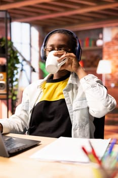 Freelancer at home wearing headphones, drinking coffee while using laptop to work on tasks. Remote worker listening music and enjoying beverage while using notebook to do job online