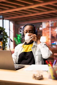 Woman drinking coffee, enjoying watching TV show during relaxation time at home. Person sitting at desk wearing headphones entertained by engrossing television series on laptop